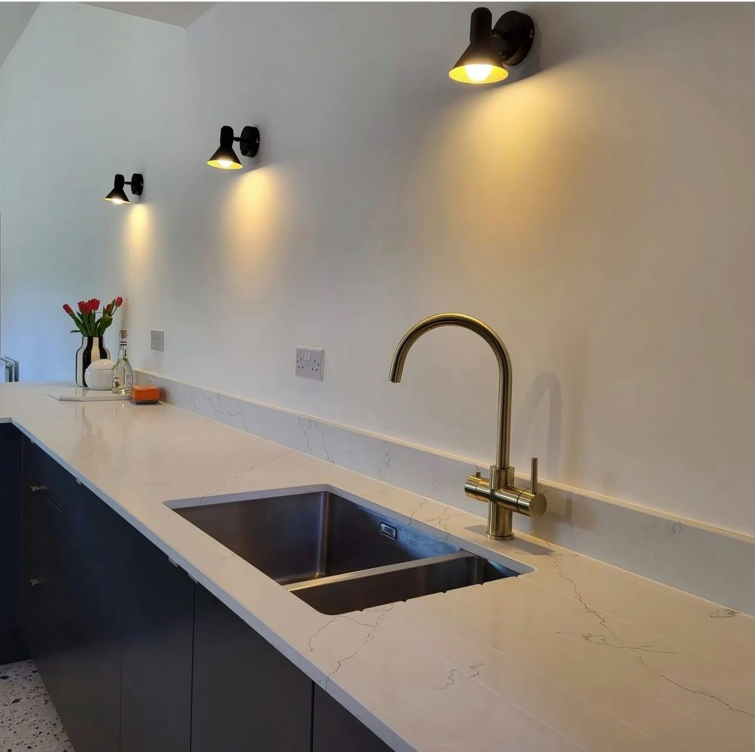 Undermount sink and brass swan-neck tap in a long Alaska run, the soft grey veining lit by the wall lights