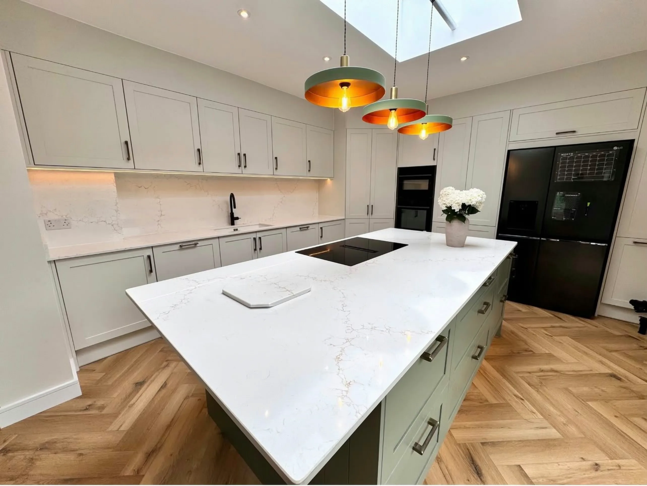 An Aurora Gold quartz island on sage green units, with the matching splashback, copper pendants and a herringbone floor, fitted by Worktop Worx