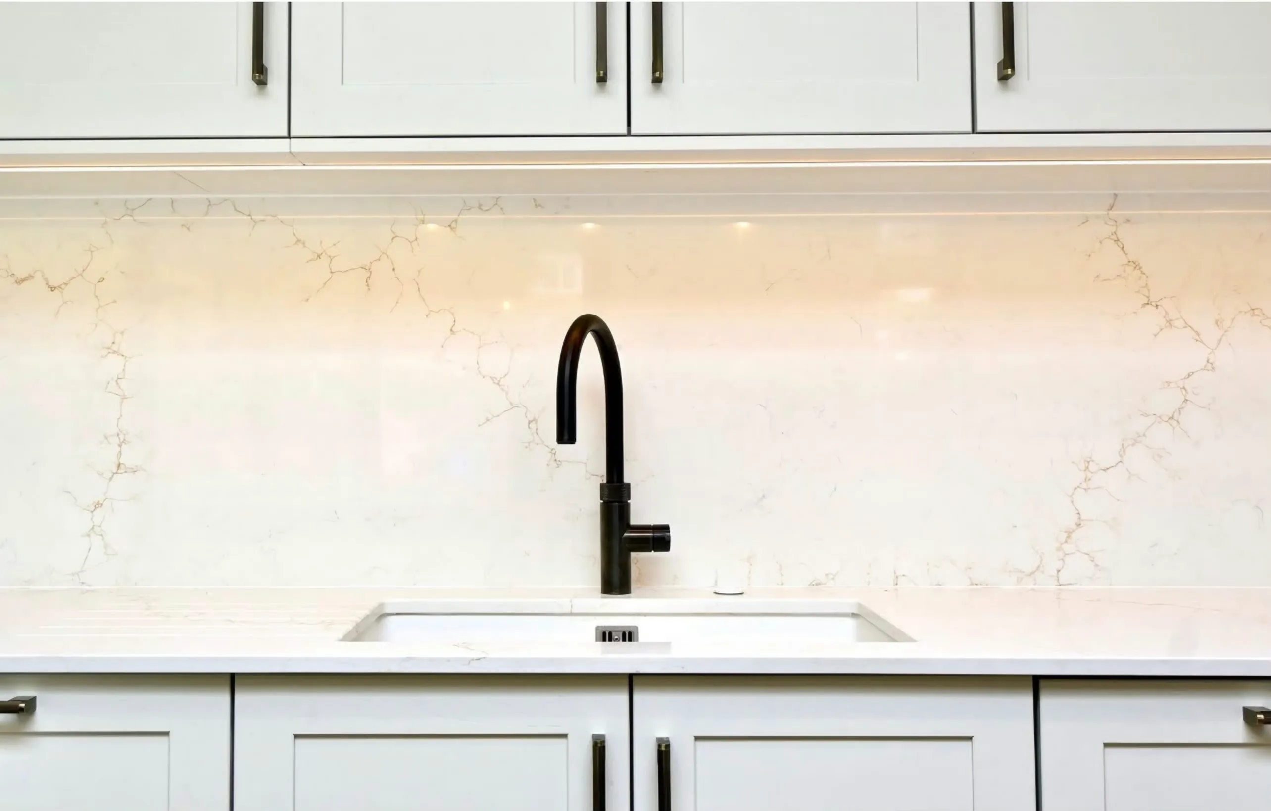 A black tap and undermount sink below a full-width Aurora Gold quartz splashback, its fine gold veining running between white shaker units