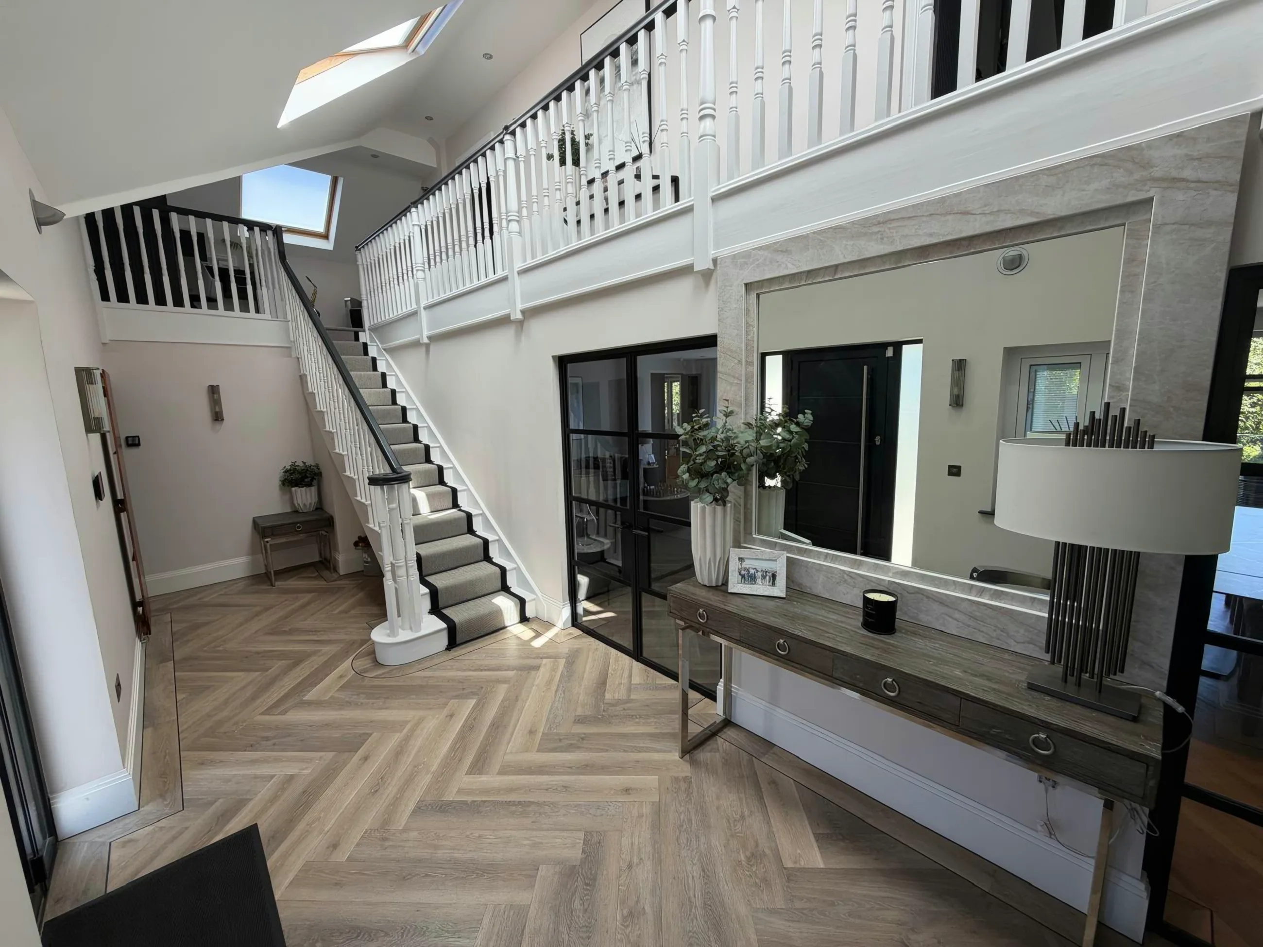 A large mirror framed in polished stone above a console table, in a double-height hallway with a herringbone floor