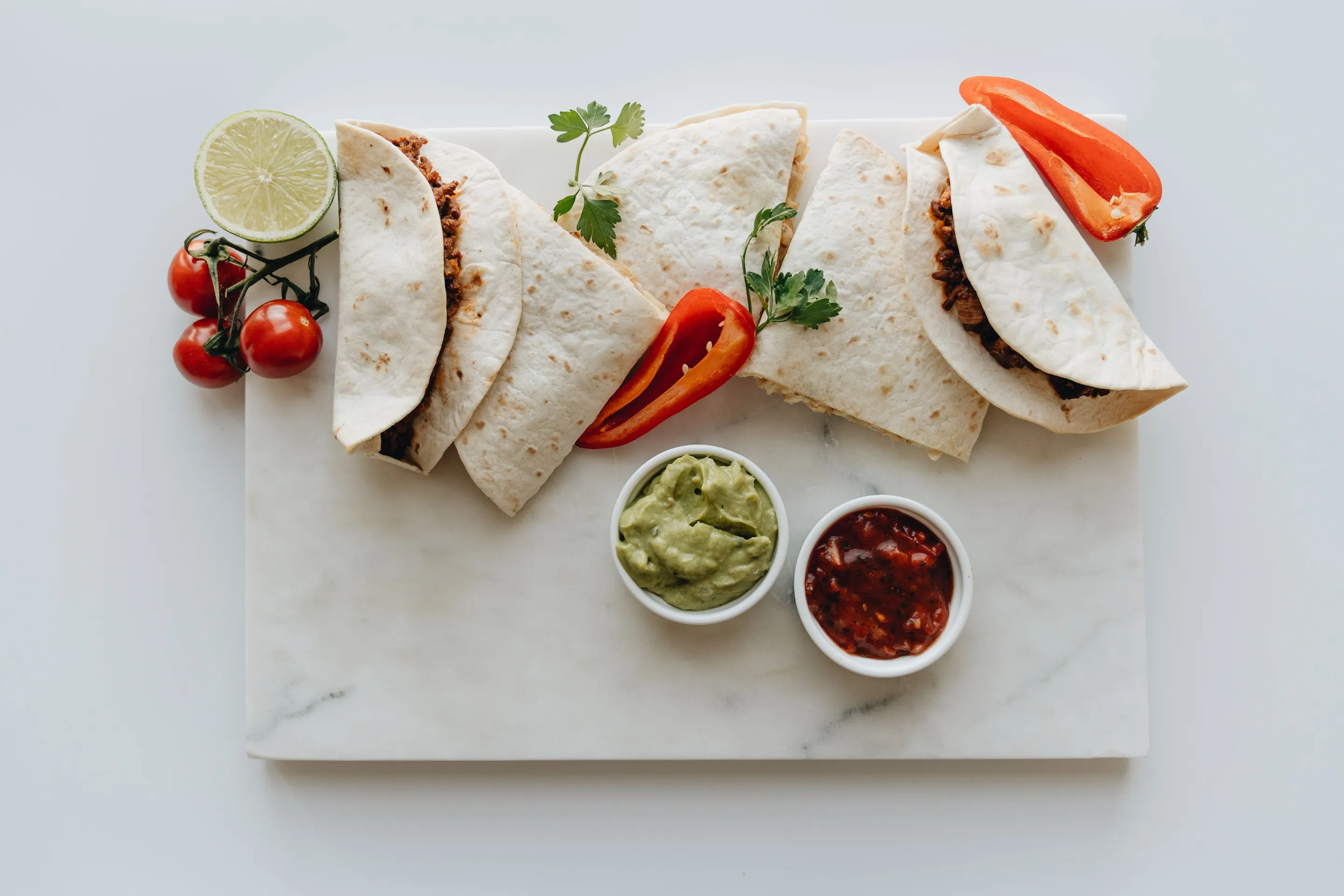 Folded tacos with guacamole and salsa on a rectangular white marble serving board