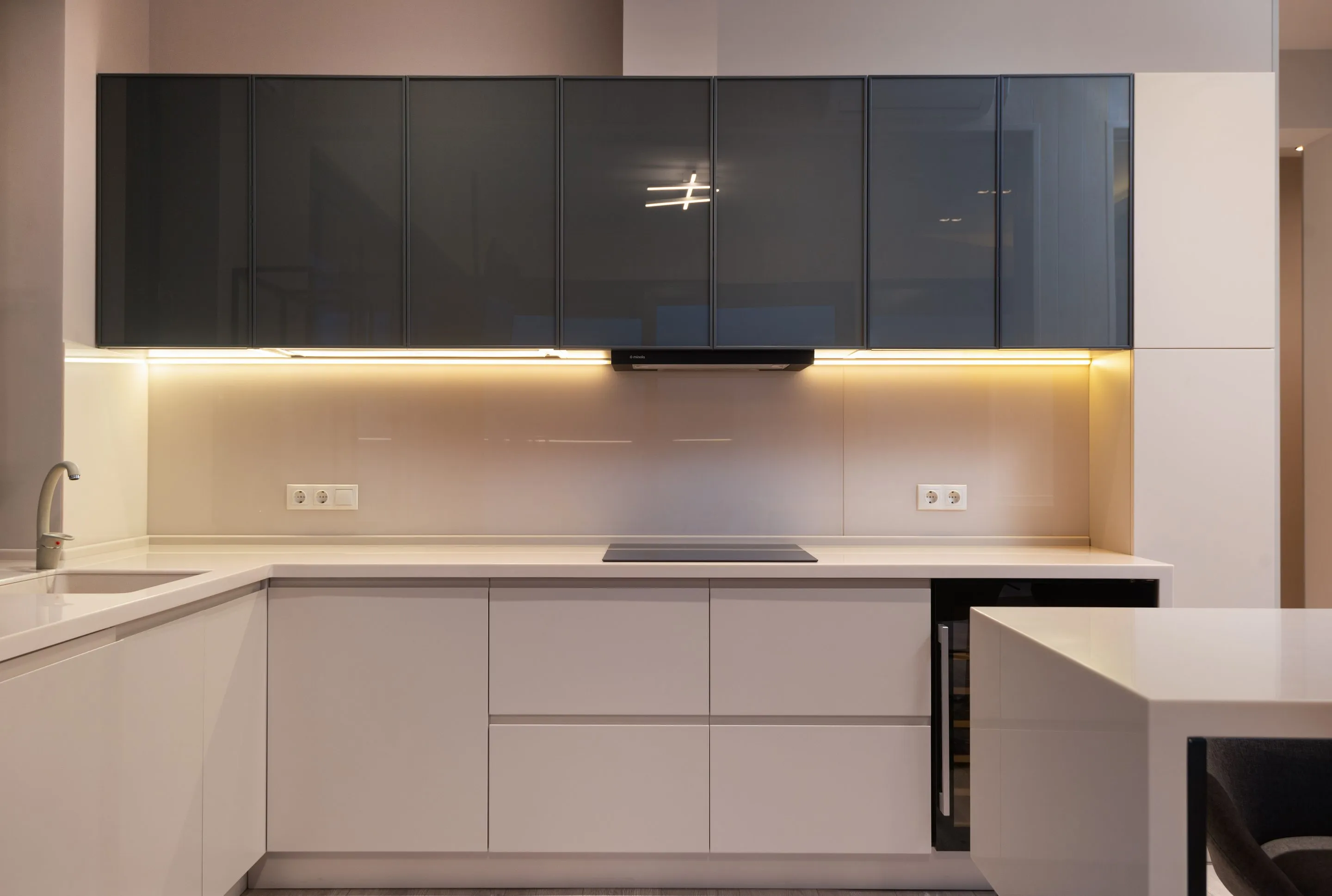 A taupe glass splashback with two sockets, lit by under-cabinet lights between dark glass wall units and a pale worktop