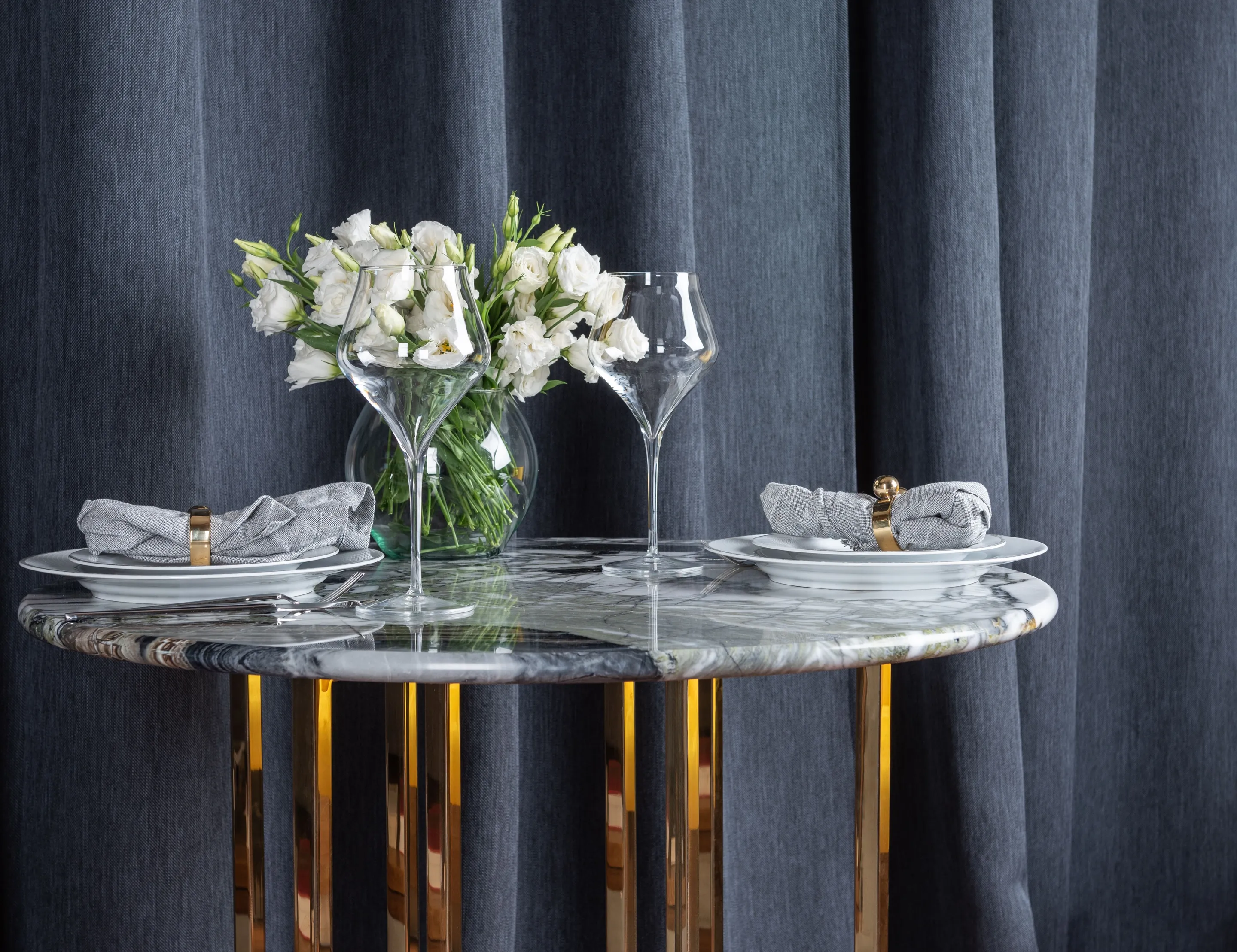 A round marble table top on a brass base, laid with plates, wine glasses and white flowers